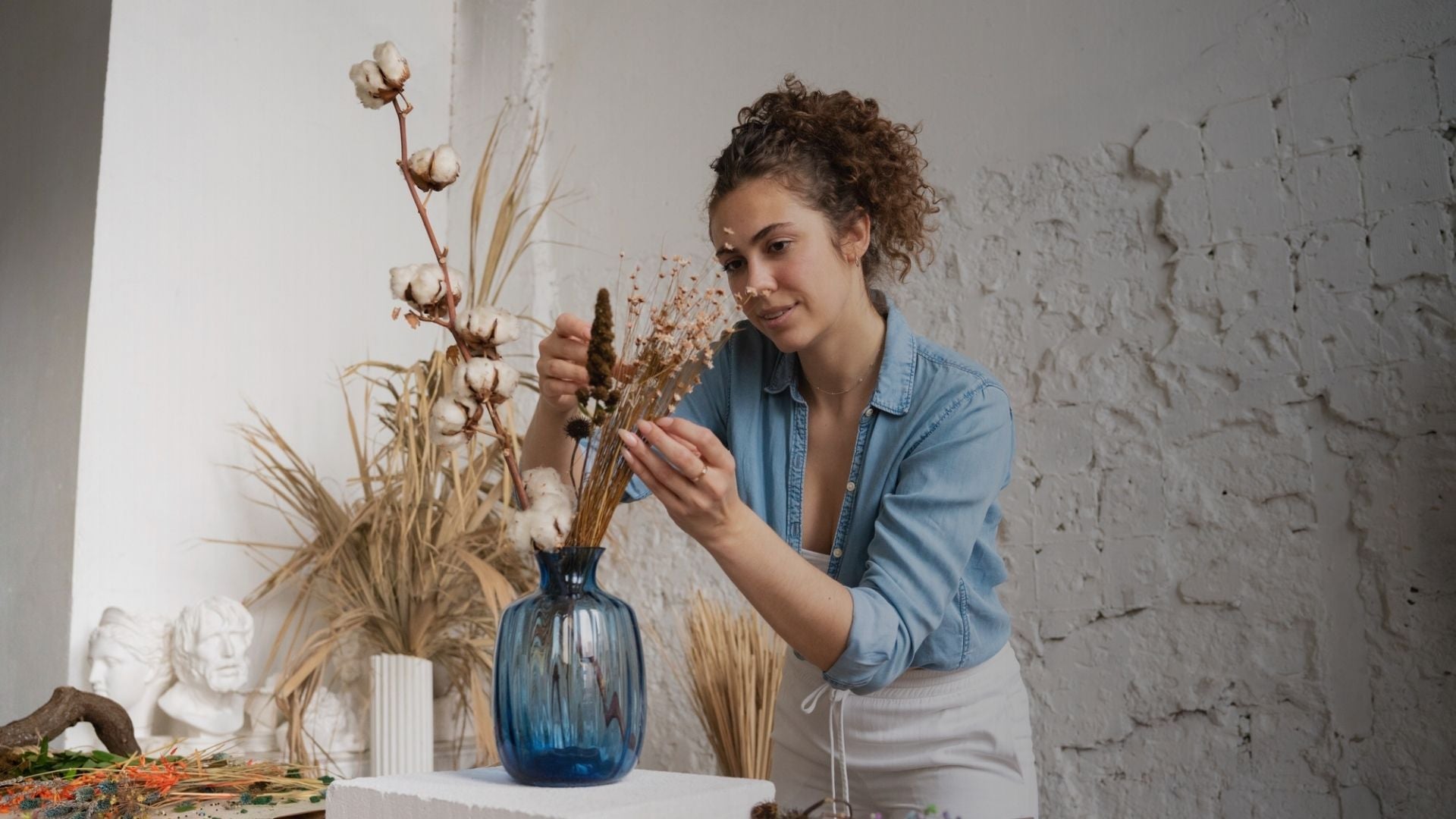 Woman arranging dried plants in a blue vase against a textured white wall.